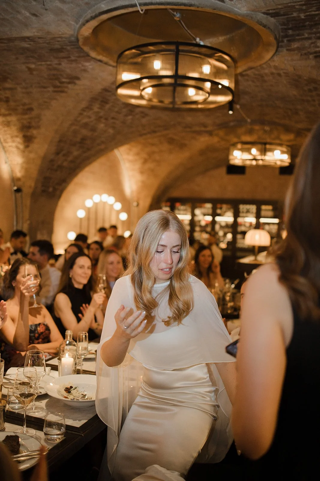 A woman in a white dress at a dinner party in a rustic, brick-ceiling room with warm lighting. She appears emotional or surprised, with other guests clapping and smiling in the background.