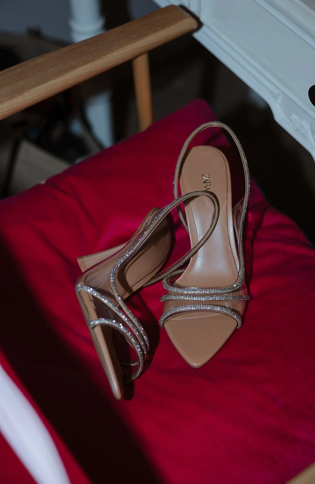A pair of beige high-heeled sandals with clear straps decorated with rhinestones, resting on a red cushioned surface.