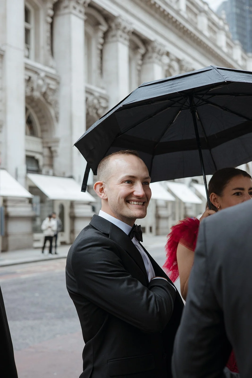 Smiling man in a tuxedo holding an umbrella at an outdoor event.