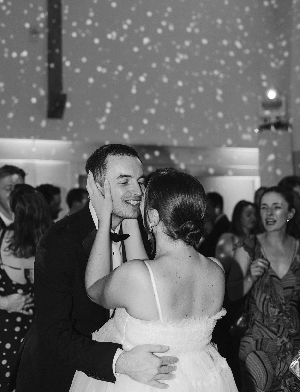 A black-and-white photo of a couple dancing closely at a formal event, with other guests in the background and decorative lighting on the ceiling.