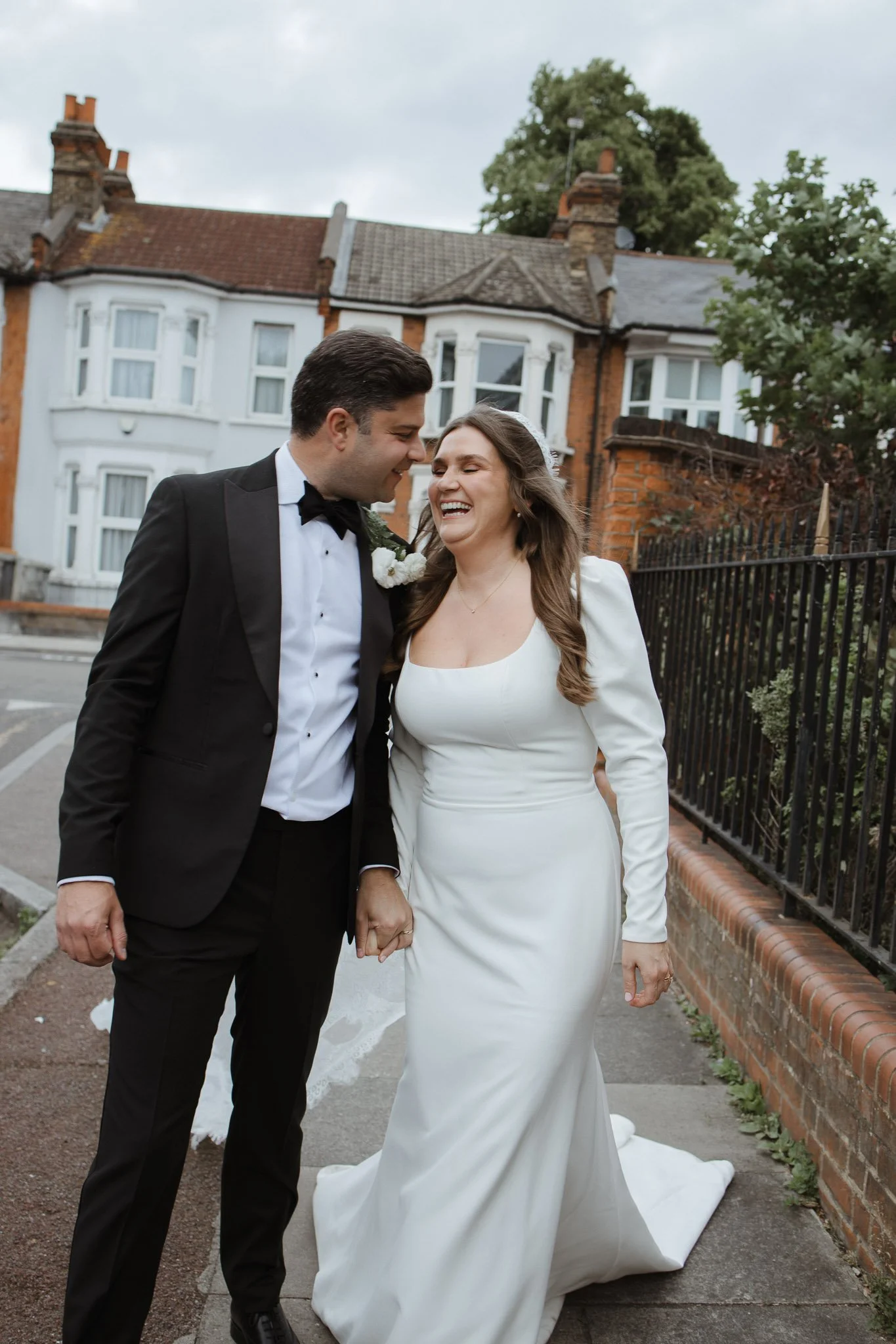 A newlywed couple dressed in wedding attire sharing a joyful moment outdoors on a sidewalk, with houses and trees in the background.