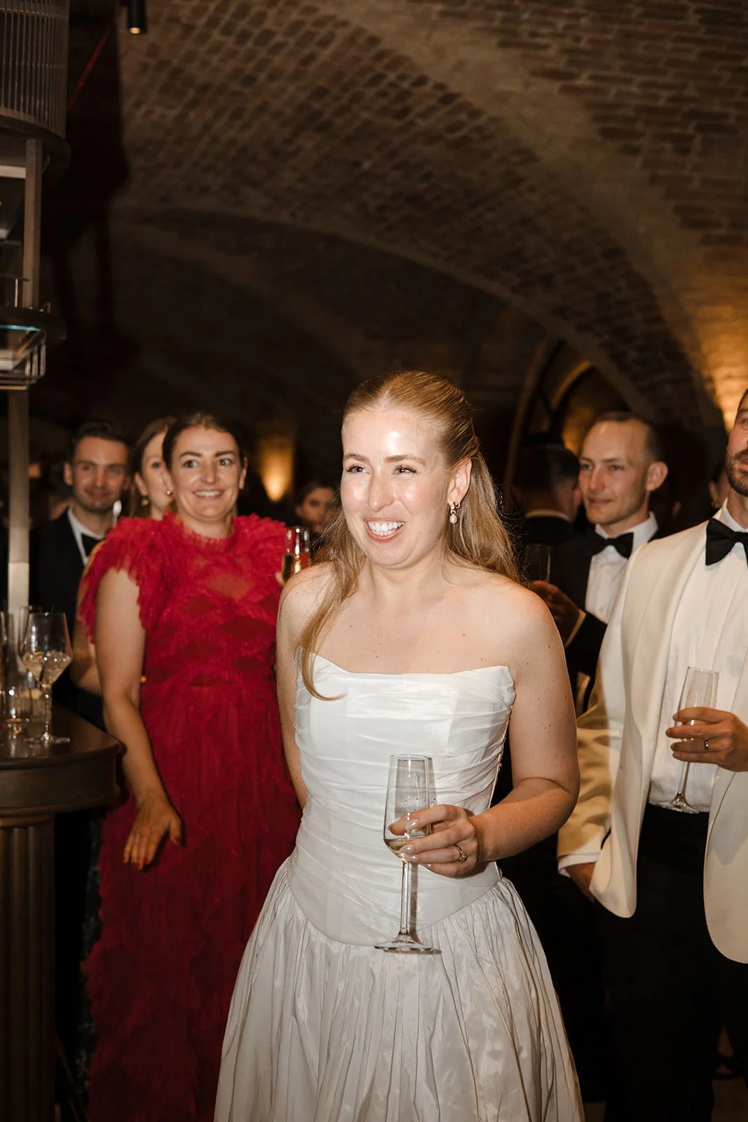 A woman in a white strapless dress holding a champagne glass, smiling at a formal event with other people dressed in tuxedos and evening gowns.