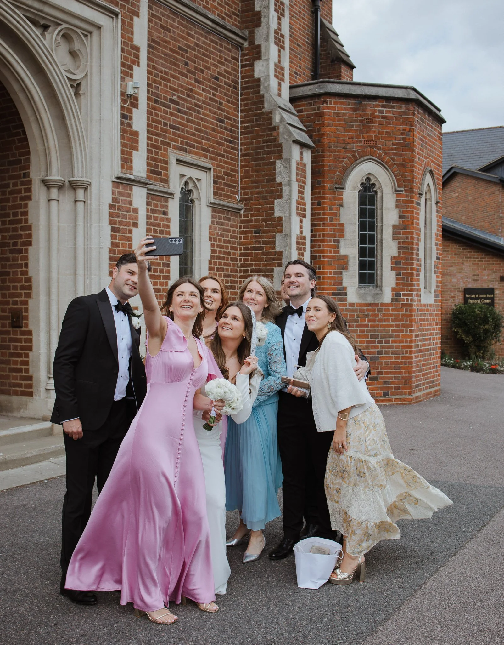 A group of seven people, dressed in formal attire, taking a selfie outside a brick church building.