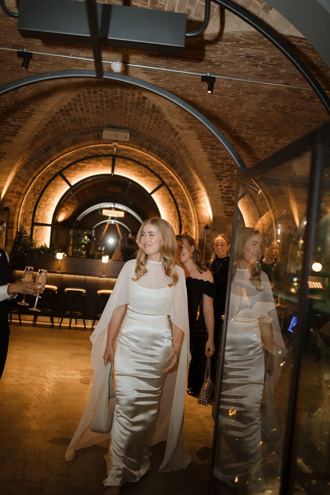 A woman in a white satin dress and cape standing in a warmly lit brick archway, smiling, with women behind her and a mirrored reflection on the right.