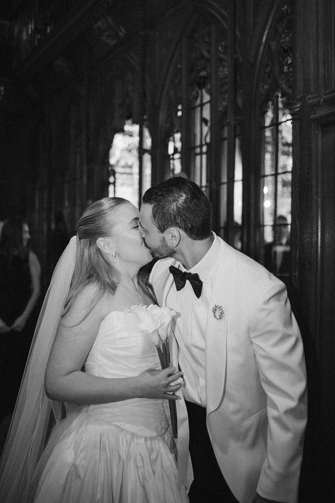 A bride and groom kiss during their wedding reception, standing near windows in an elegant, dark wood-paneled room, with the bride holding a bouquet of white calla lilies.