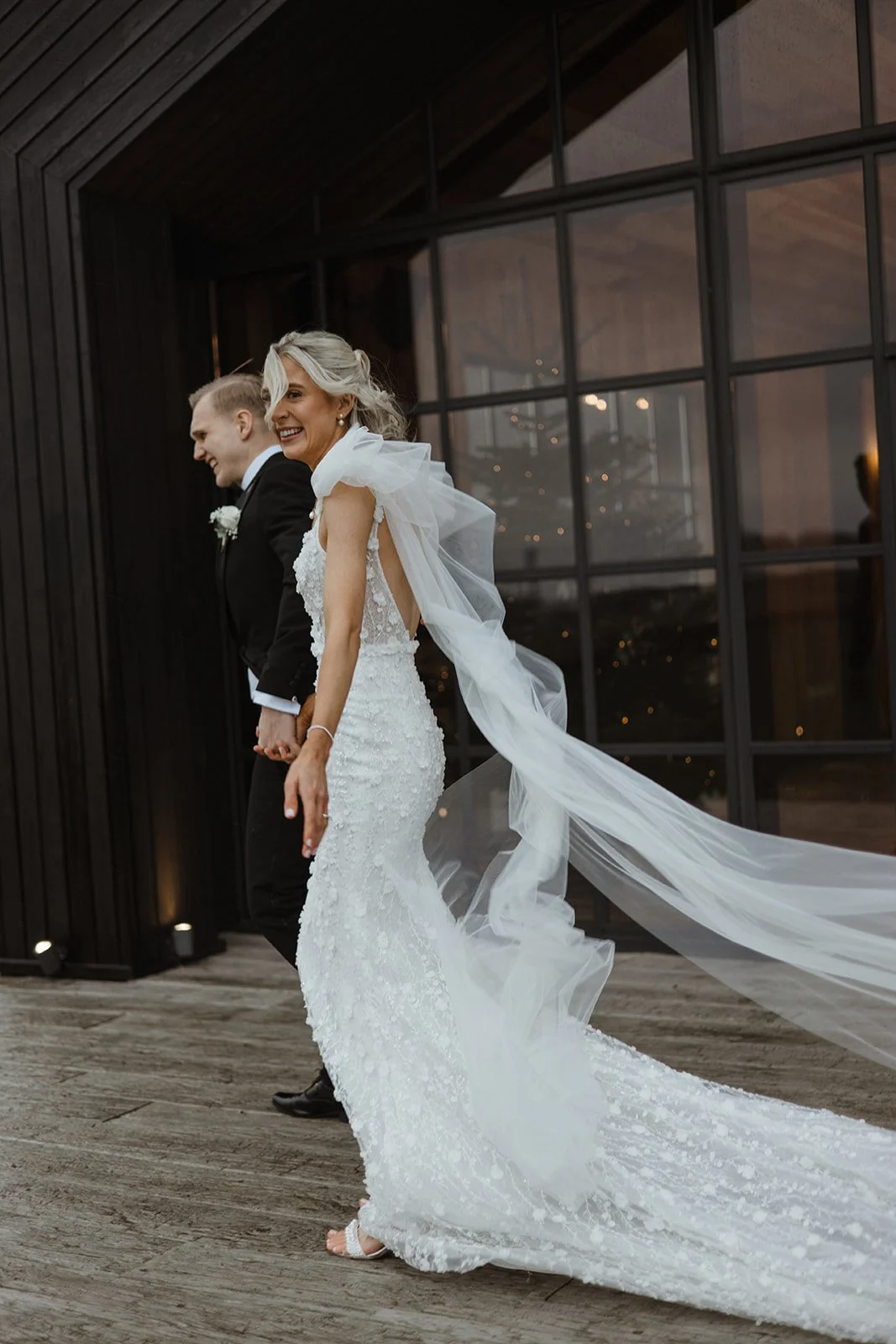 Bride and groom holding hands, walking and smiling during sunset outside a modern building.