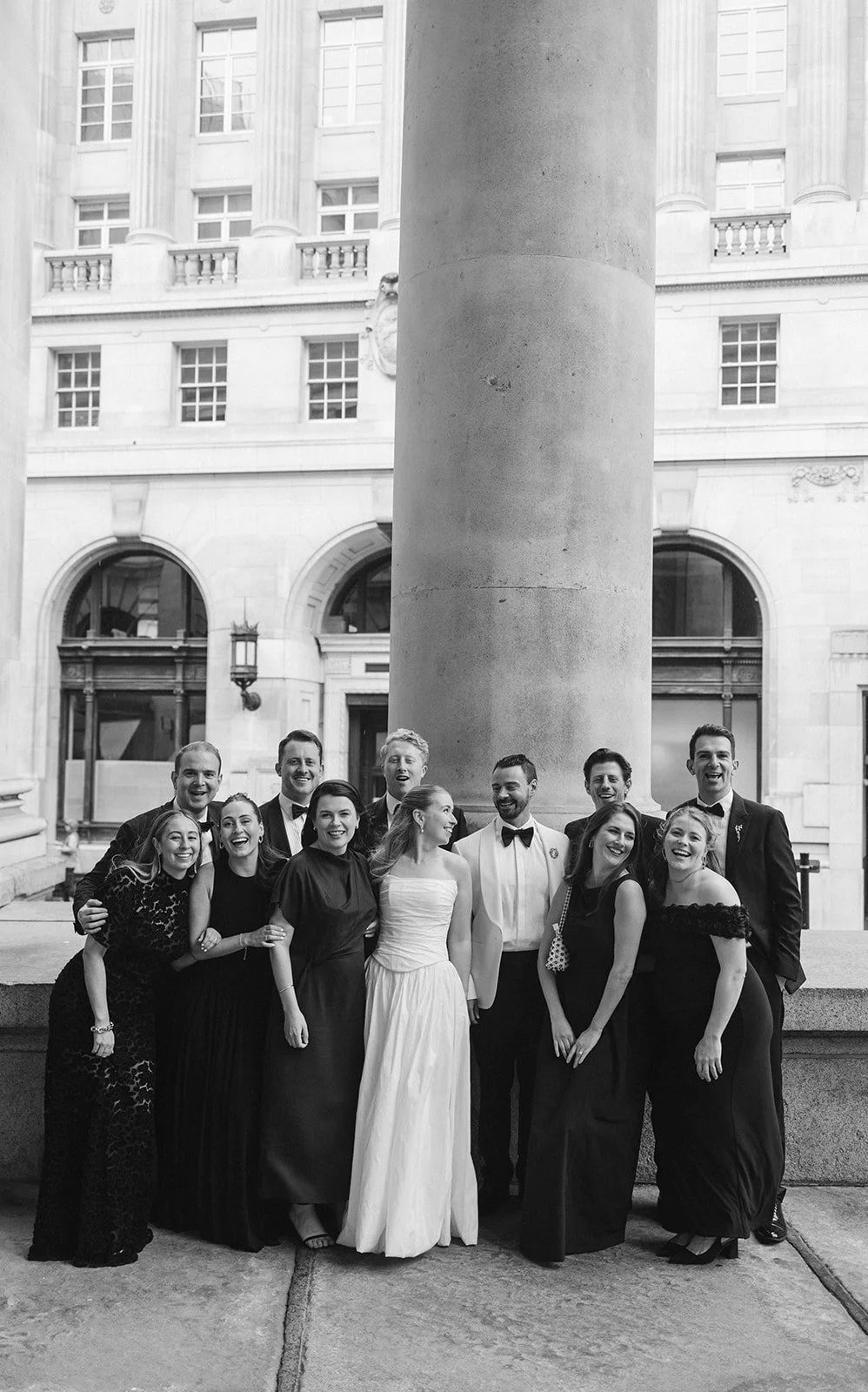 A group of people dressed in formal attire, gathered outdoors in front of a large column with a building in the background, smiling and posing for a photo.