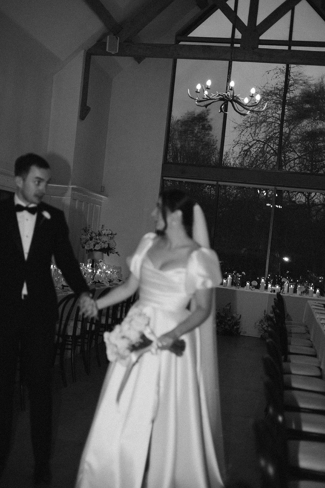 A black and white photo of a bride and groom holding hands at a wedding reception. The bride is wearing a white gown and veil, holding a bouquet of flowers, while the groom is in a tuxedo. They are inside a decorated venue with large windows and a ch