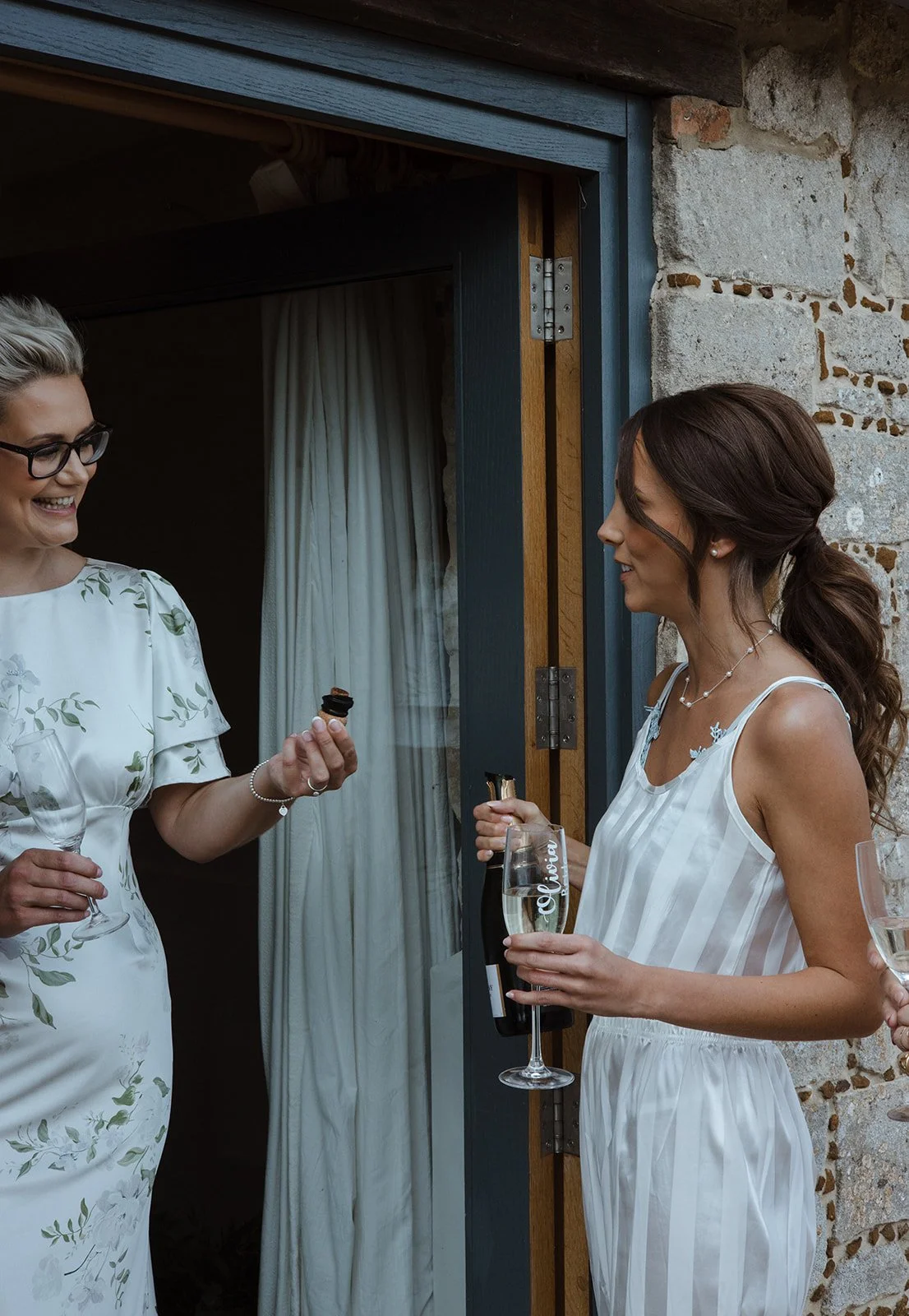 Two women in white dresses sharing a toast with champagne and a woman holding a small snack, standing near a stone wall and a doorway with a curtain inside.