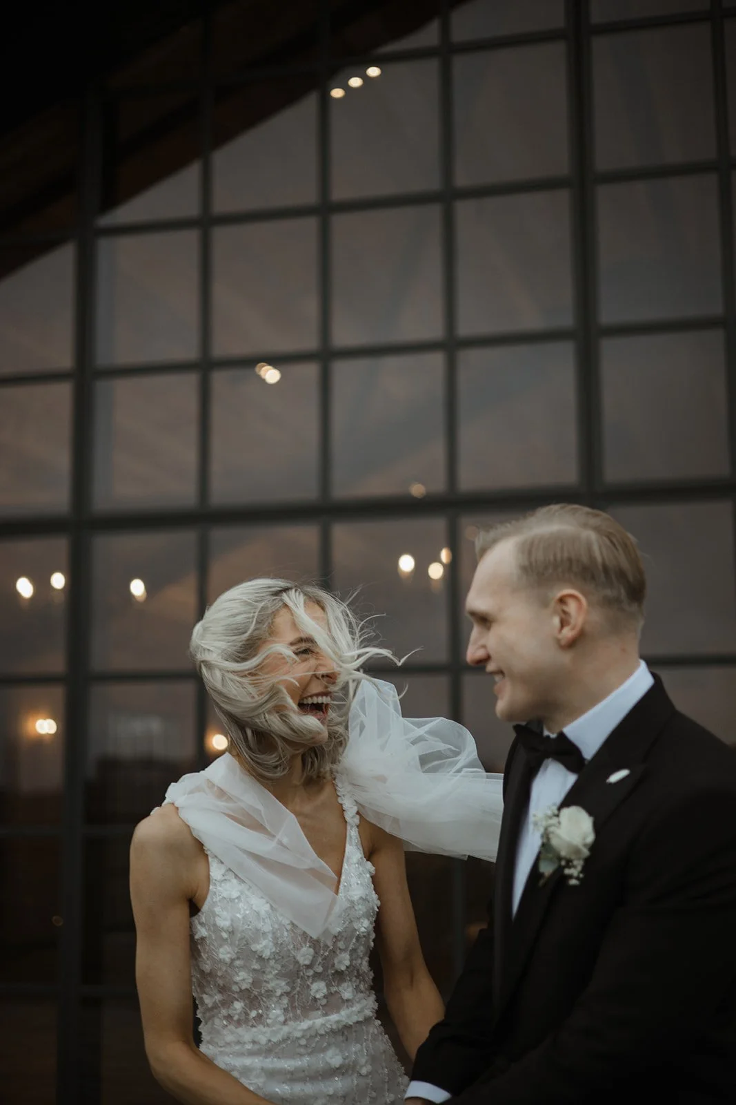 A bride and groom are smiling and laughing during their wedding reception, standing in front of a large glass window with reflections of lights.