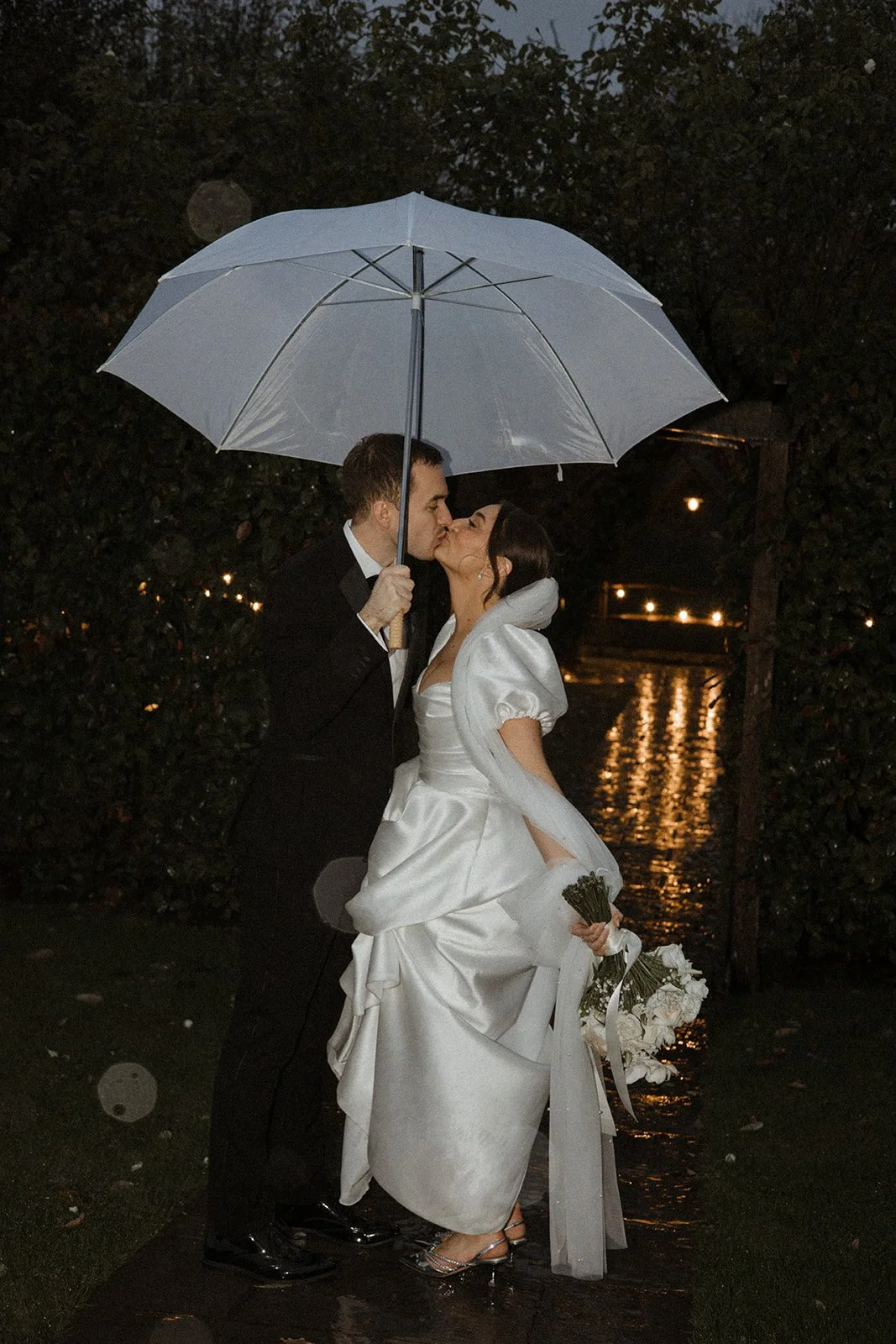 A newlywed couple sharing a kiss under an umbrella during a rainy night, standing on a wet pathway near a body of water, with the bride holding a bouquet of white flowers.