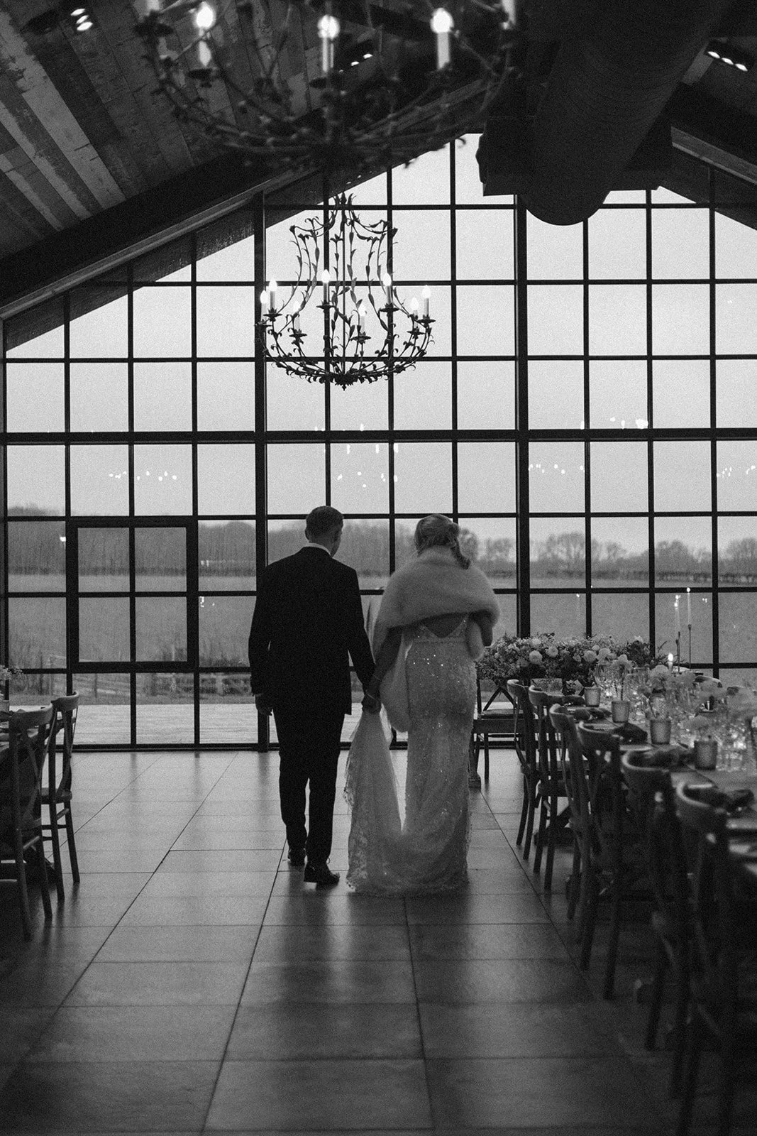 A couple walking hand in hand inside a large, modern venue with tall windows and a chandelier hanging from the ceiling, possibly at a wedding reception.
