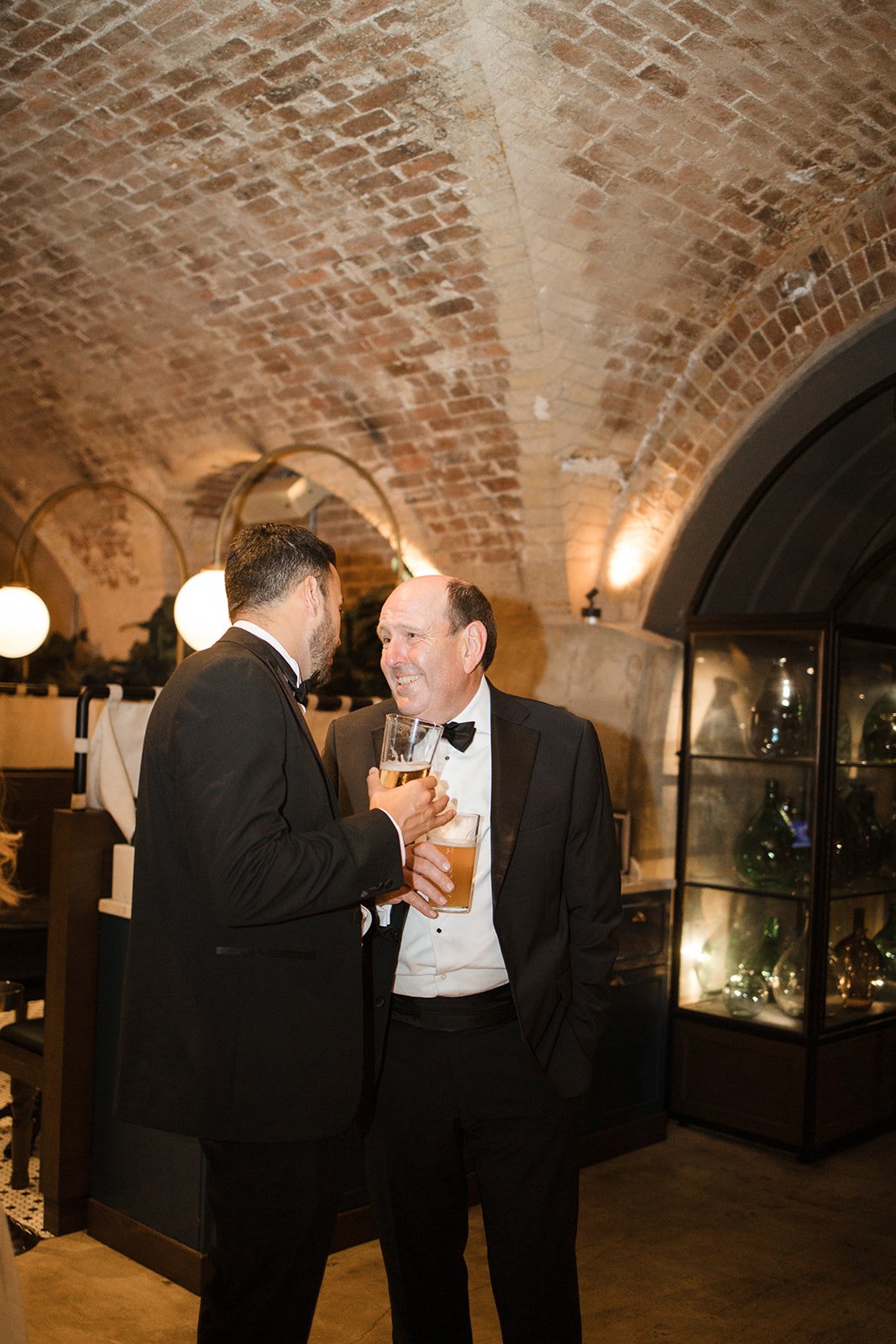 Two men in tuxedos talking and holding drinks in a brick arched room with warm lighting.