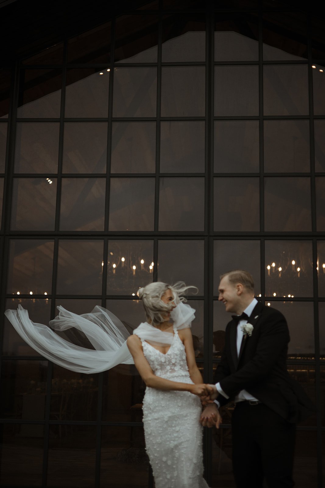 A bride and groom holding hands and smiling at each other in front of large glass windows at a wedding reception or ceremony, with interior chandelier lighting visible through the windows.