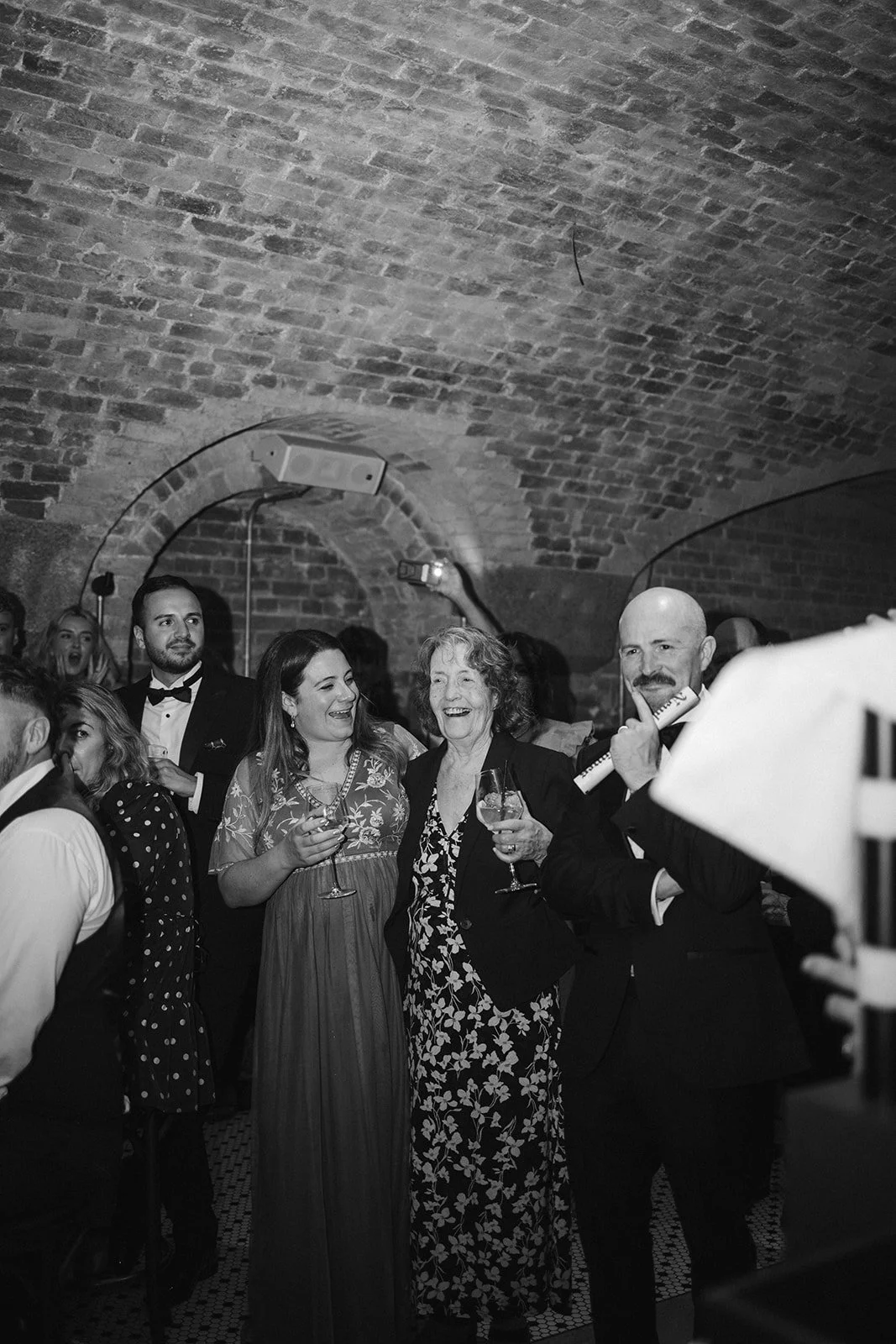 People smiling and celebrating at a party or event in a brick-walled venue, with some holding glasses of wine and using a musical instrument as a prop.