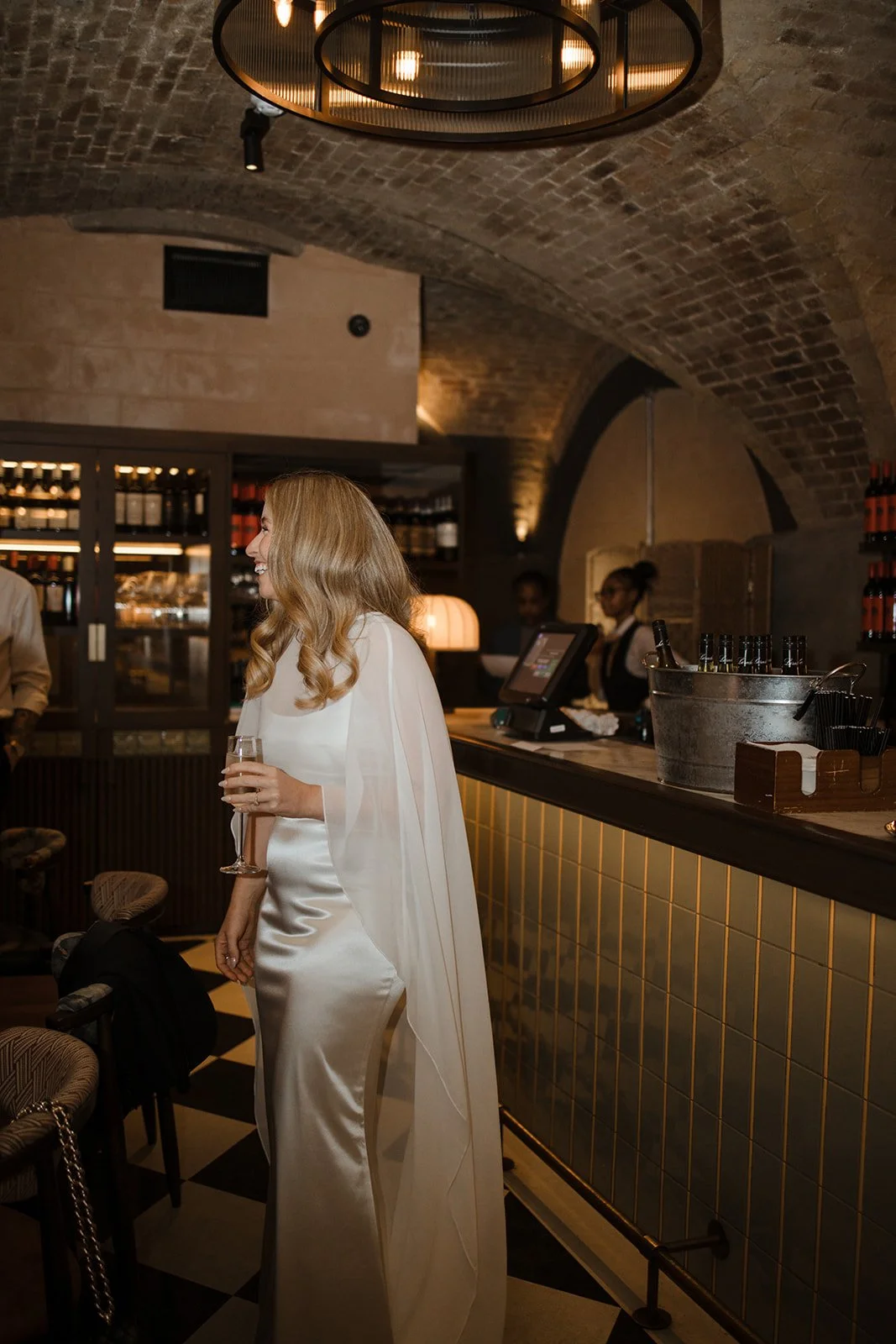 A woman in a silky white gown holding a glass of champagne at a bar with a rustic brick arched ceiling, and bar staff working behind the counter.