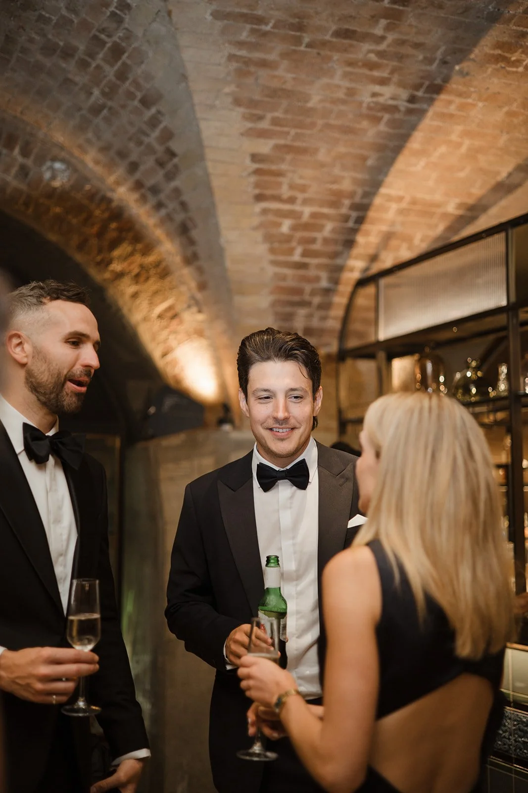 Three men and one woman in formal attire talking and holding drinks at a social event in a brick-arched room.