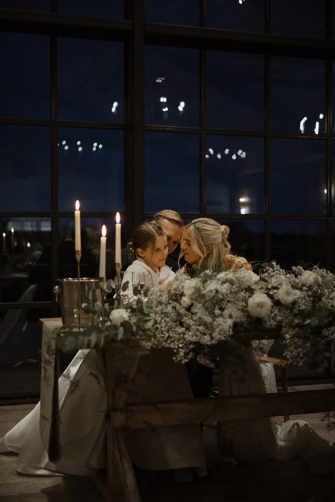 A family of three, including a woman, a man, and a young girl, sit closely at a decorated table during a nighttime celebration, with candles and flowers.