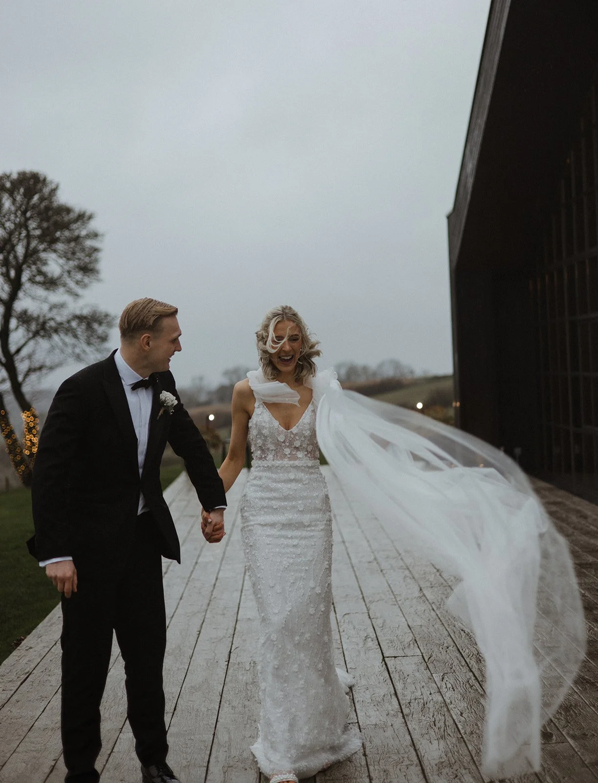 Bride and groom holding hands, walking on a wooden pathway outdoors, bride in a lace wedding dress with a long veil, groom in a tuxedo, overcast sky.