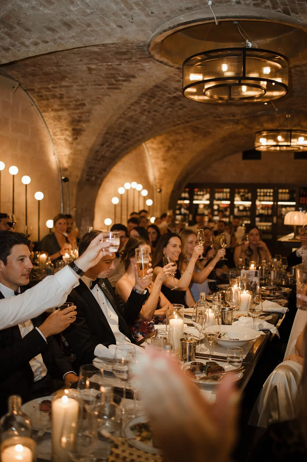 Guests at a formal dinner in a wine cellar-like setting raising glasses in a toast, with candles and elegant table settings.