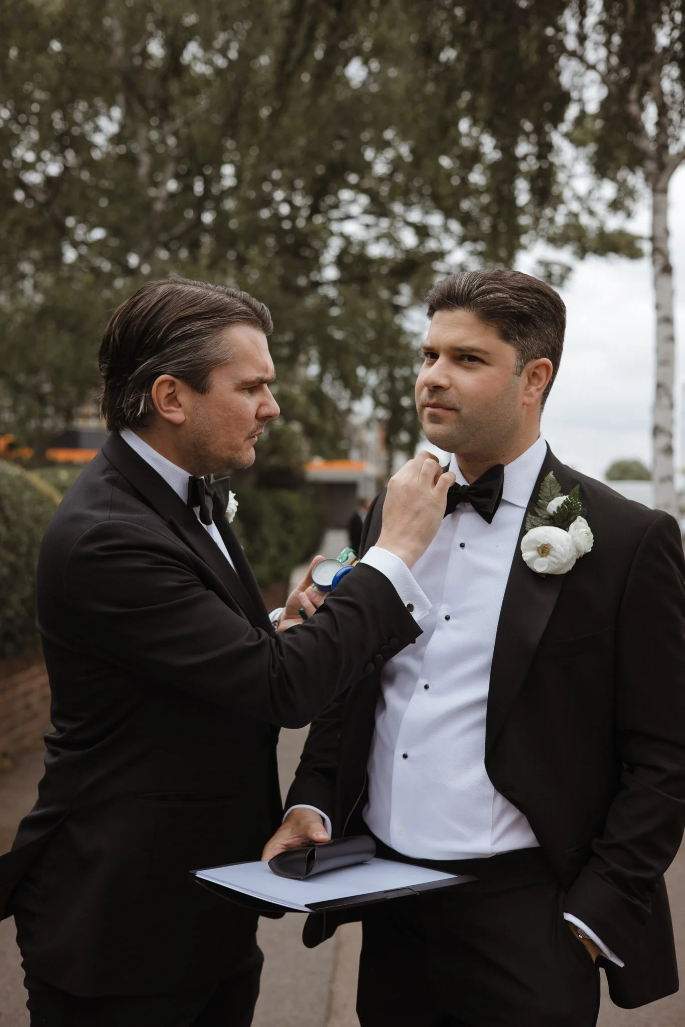 Two men in tuxedos, one adjusting the other's bow tie during an outdoor wedding or formal event.