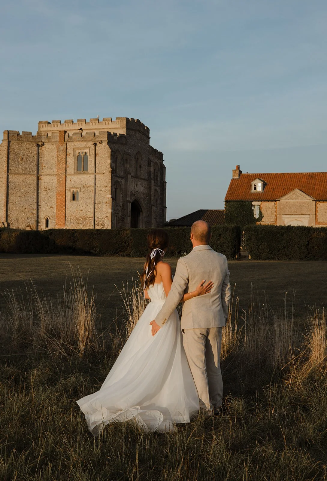 A bride and groom walking in a field at sunset with a castle in the background, dressed in wedding attire.