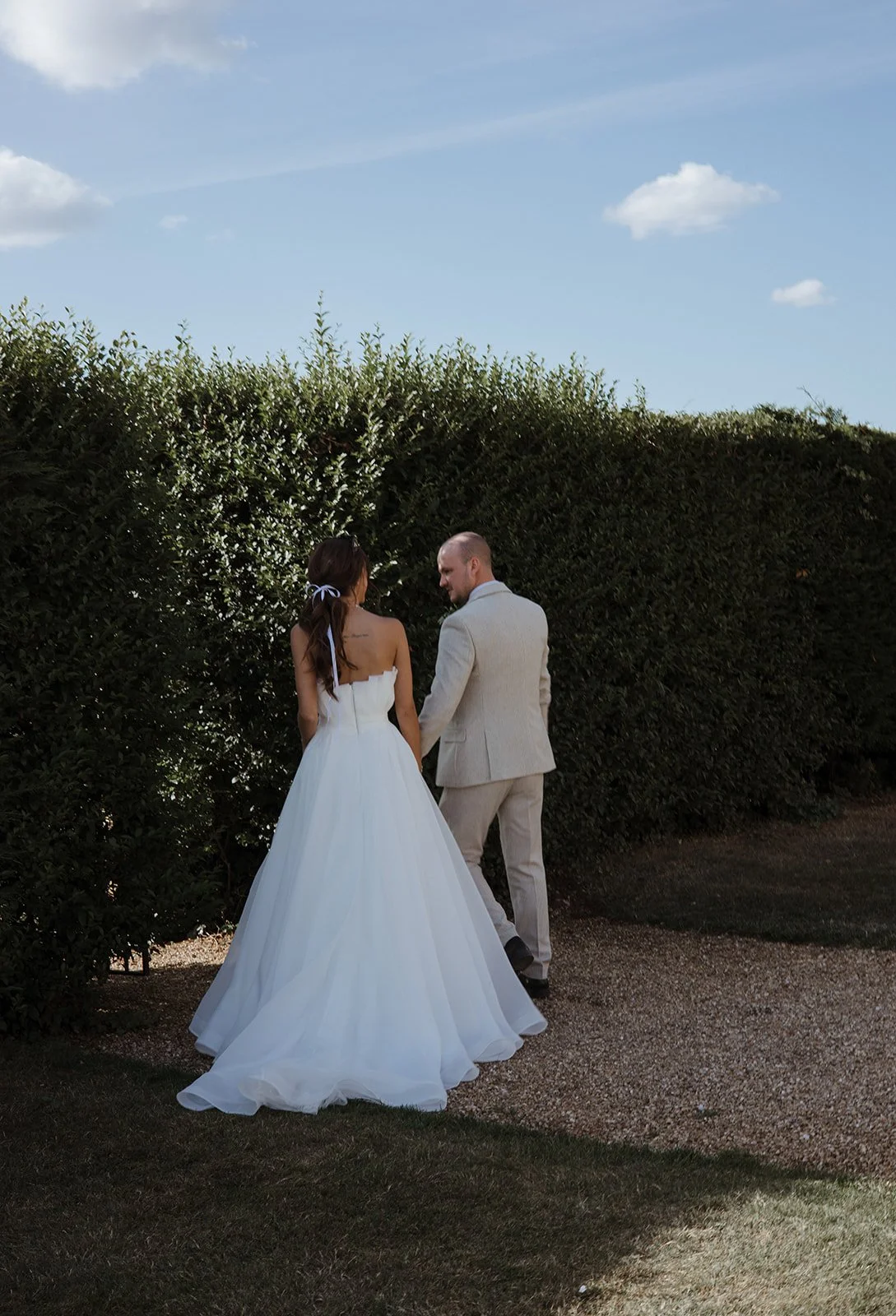 A bride in a white wedding dress and a groom in a light-colored suit are walking together outdoors, holding hands, near a tall hedge on a sunny day.