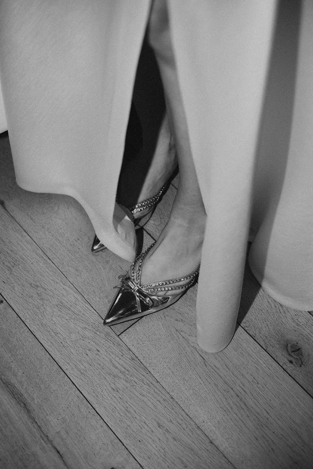 Black and white photo of a person's feet wearing pointed high-heeled shoes with decorative straps and bows, partially covered by a dress or skirt.