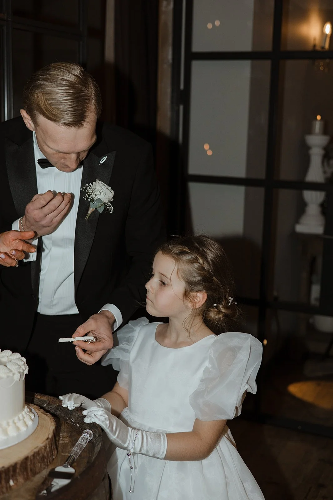 A young girl in a white dress and gloves at a wedding cake table, with an older man in a tuxedo, possibly a groom, observing her. The girl appears to be preparing to cut or serve the cake.