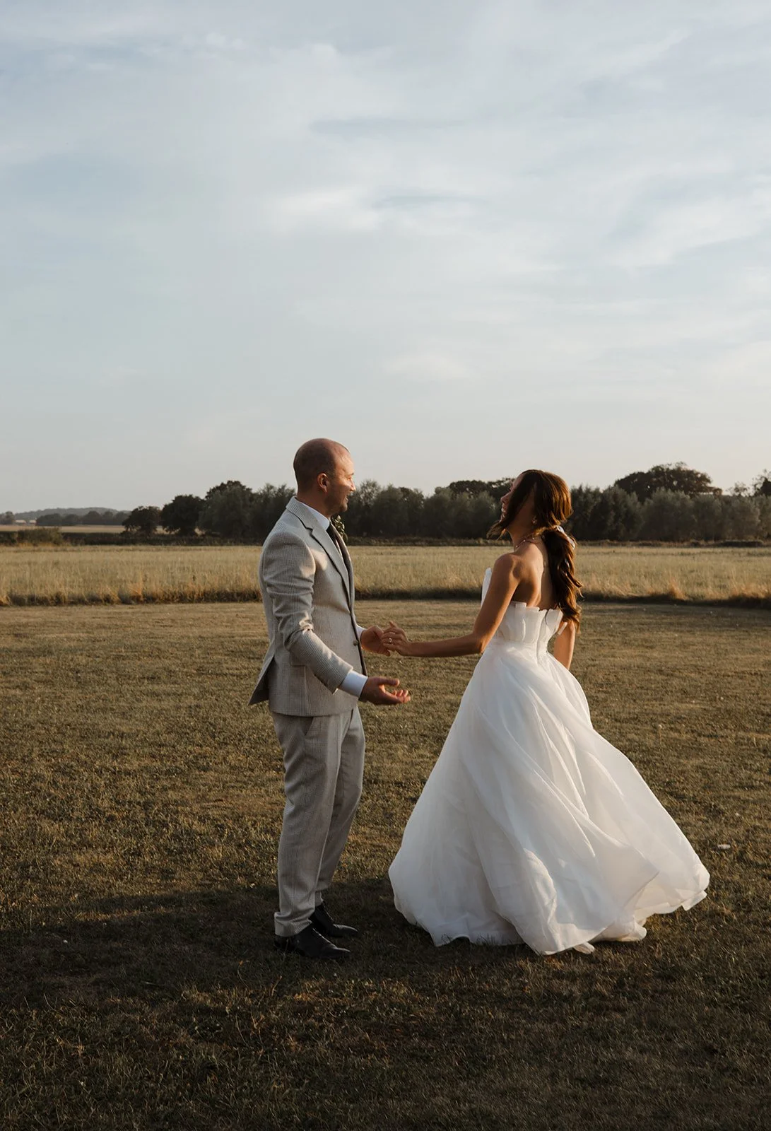 A bride and groom holding hands in a field during sunset, with open sky and distant trees in the background.