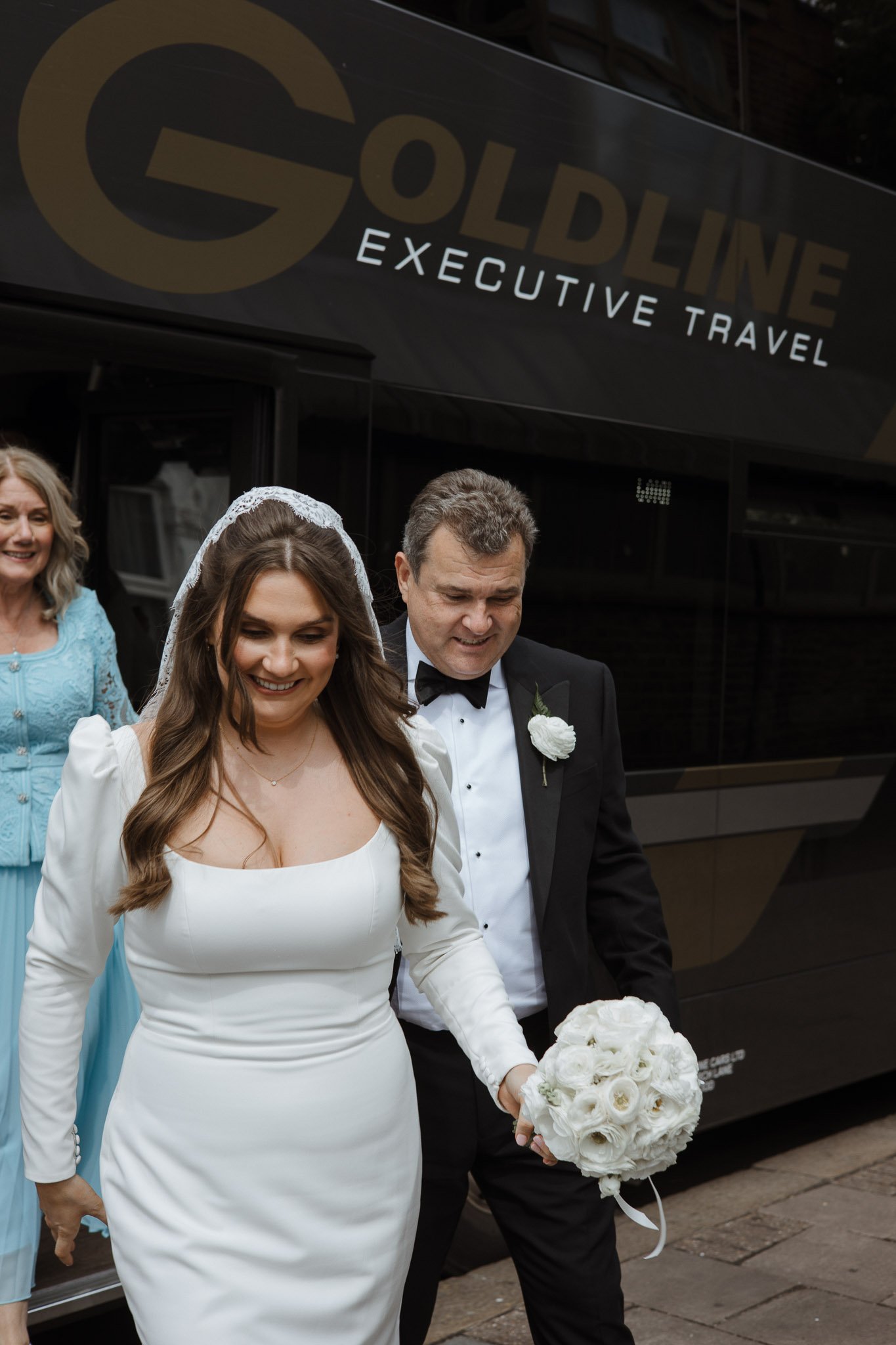 A bride and groom walking with a bouquet, smiling, after their wedding, with a woman in a blue dress in the background, outside a travel company bus.