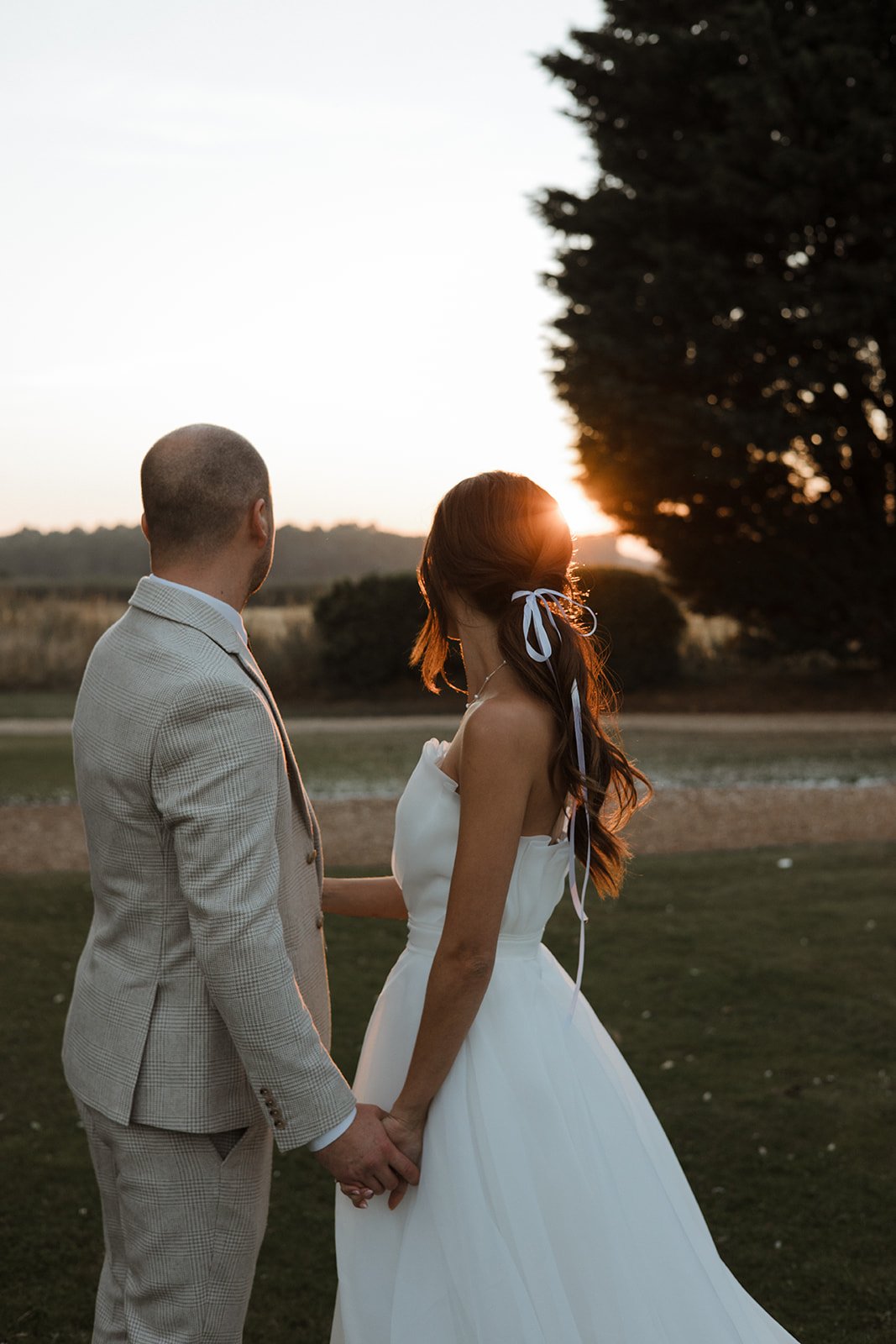 A bride and groom holding hands outdoors during sunset, with the bride wearing a white wedding dress and the groom in a light-colored suit, standing on a grassy area with trees in the background.