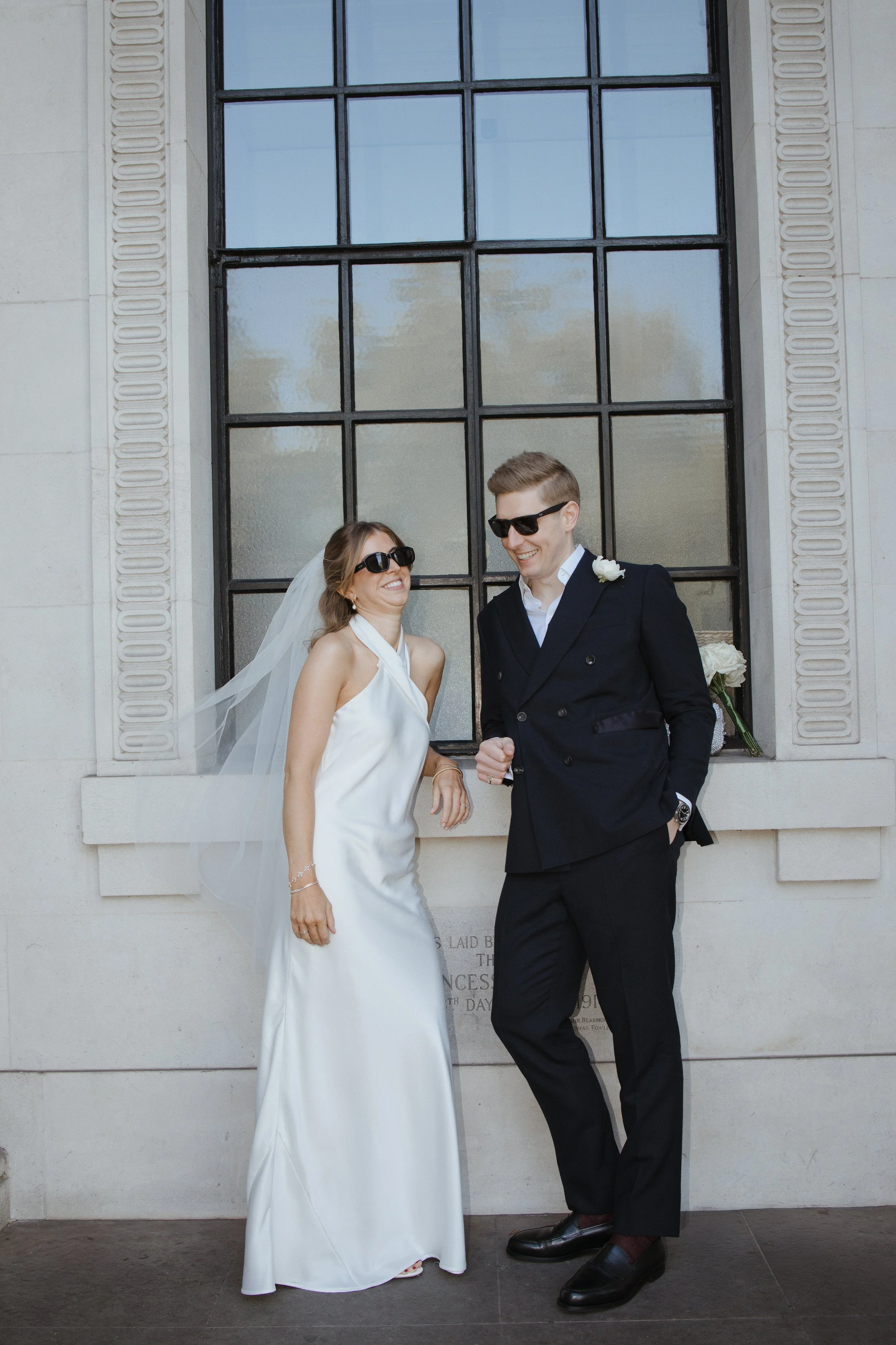 A bride and groom wearing black sunglasses, standing in front of a building with a large window. The bride is in a white wedding dress with a veil, and the groom is in a dark suit with a white shirt. They are smiling and holding hands, with flowers in the background.