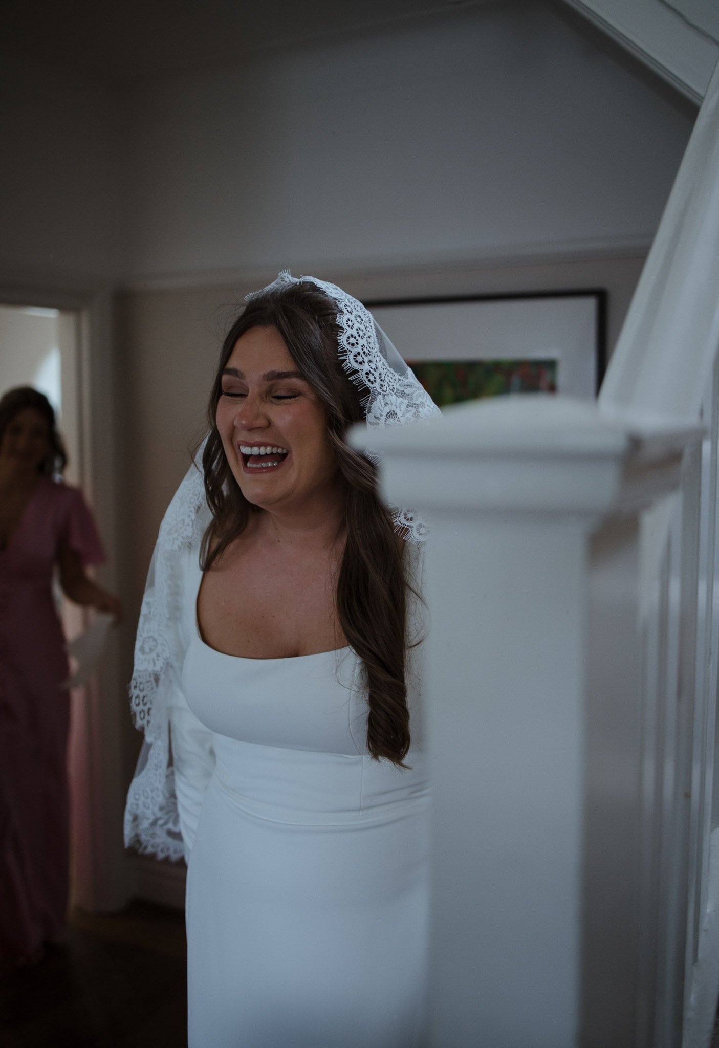 A bride in a white dress and a lace veil is smiling and laughing, standing indoors with two women in pink dresses in the background.