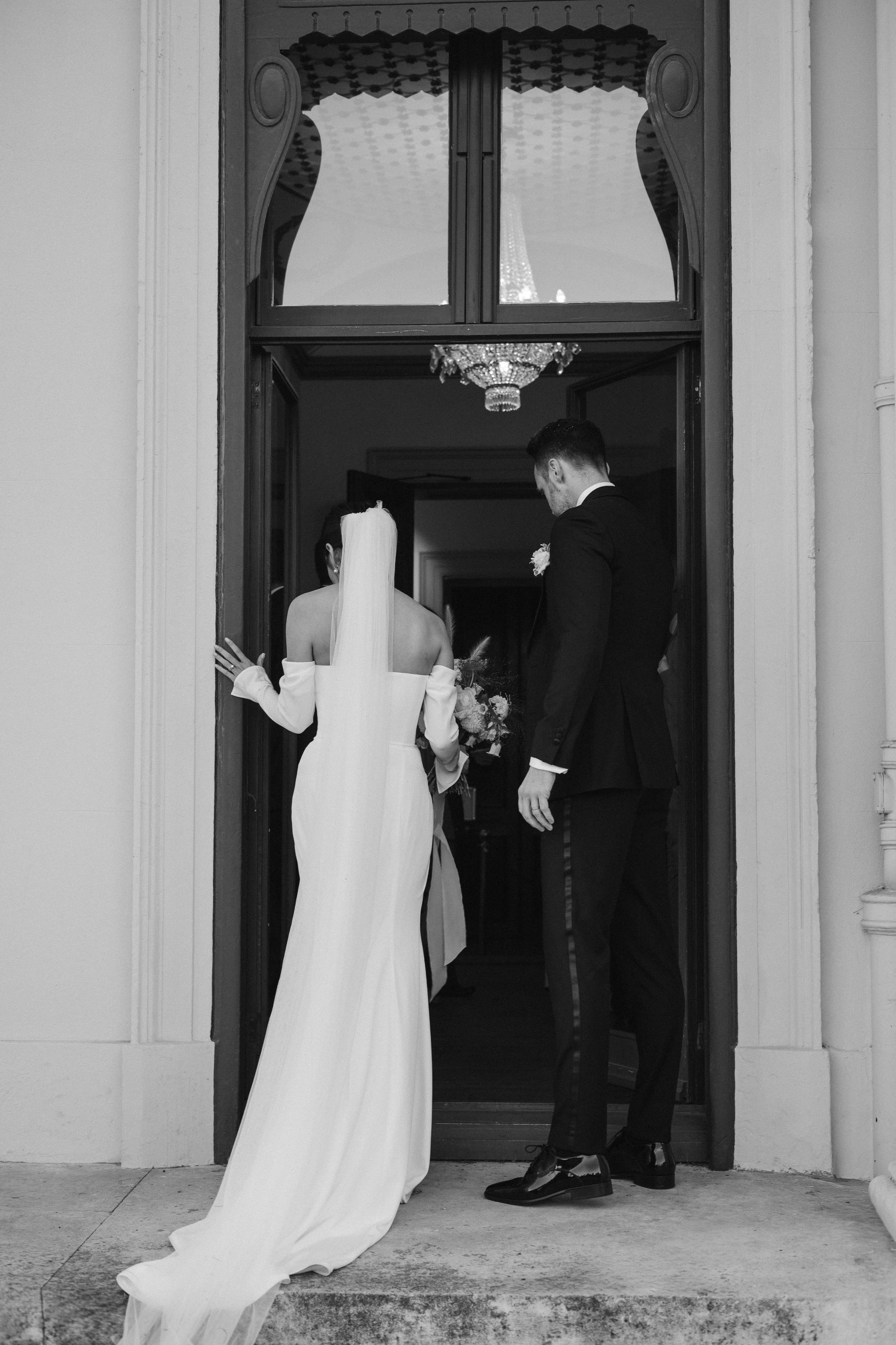 A bride and groom standing at a doorway, facing away from the camera, with a chandelier visible inside the room.