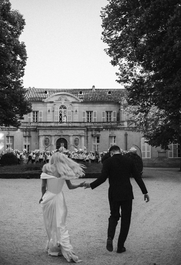 A couple, a woman in a wedding dress and a man in a suit, walking hand in hand outdoors in front of a large mansion. The scene is in black and white, with a group of people gathered in the background.