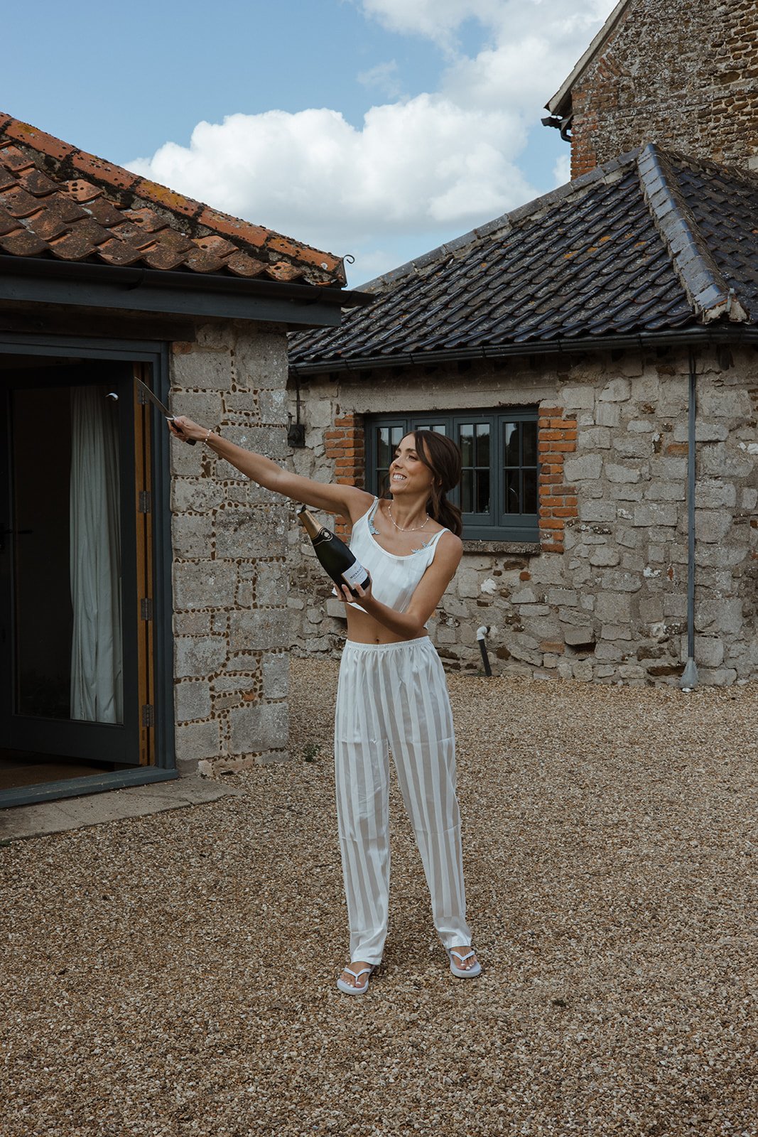A woman in white striped pajamas is celebrating outside a rustic stone house with a glass of champagne in one hand and a bottle of champagne in the other, raising her arm in a toast.