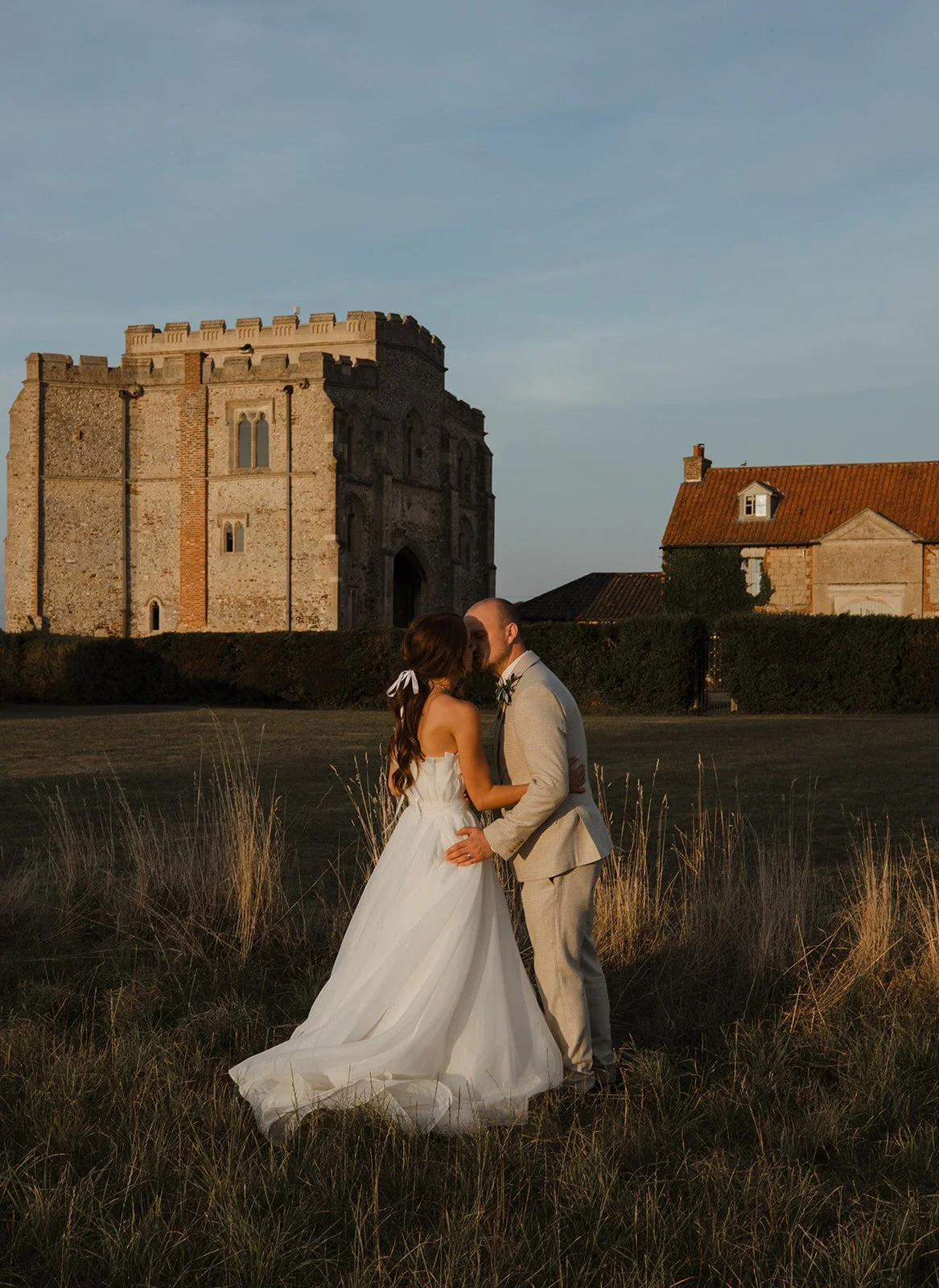 Bride and groom in wedding attire embracing in an open field with a historic castle and a stone house in the background during sunset.