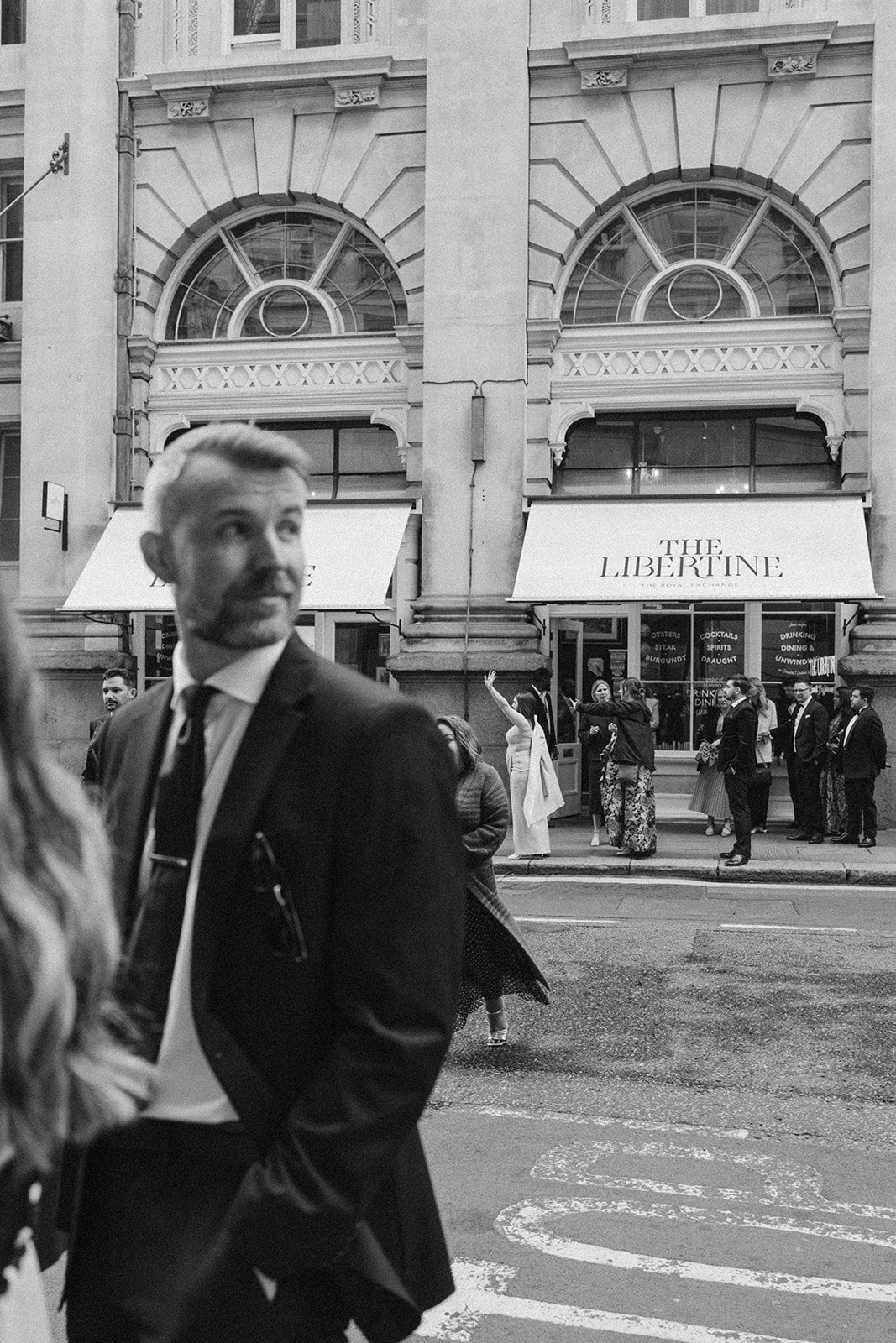 People waiting outside a building named 'The Libertine' on a city street, with a man in a suit in the foreground.