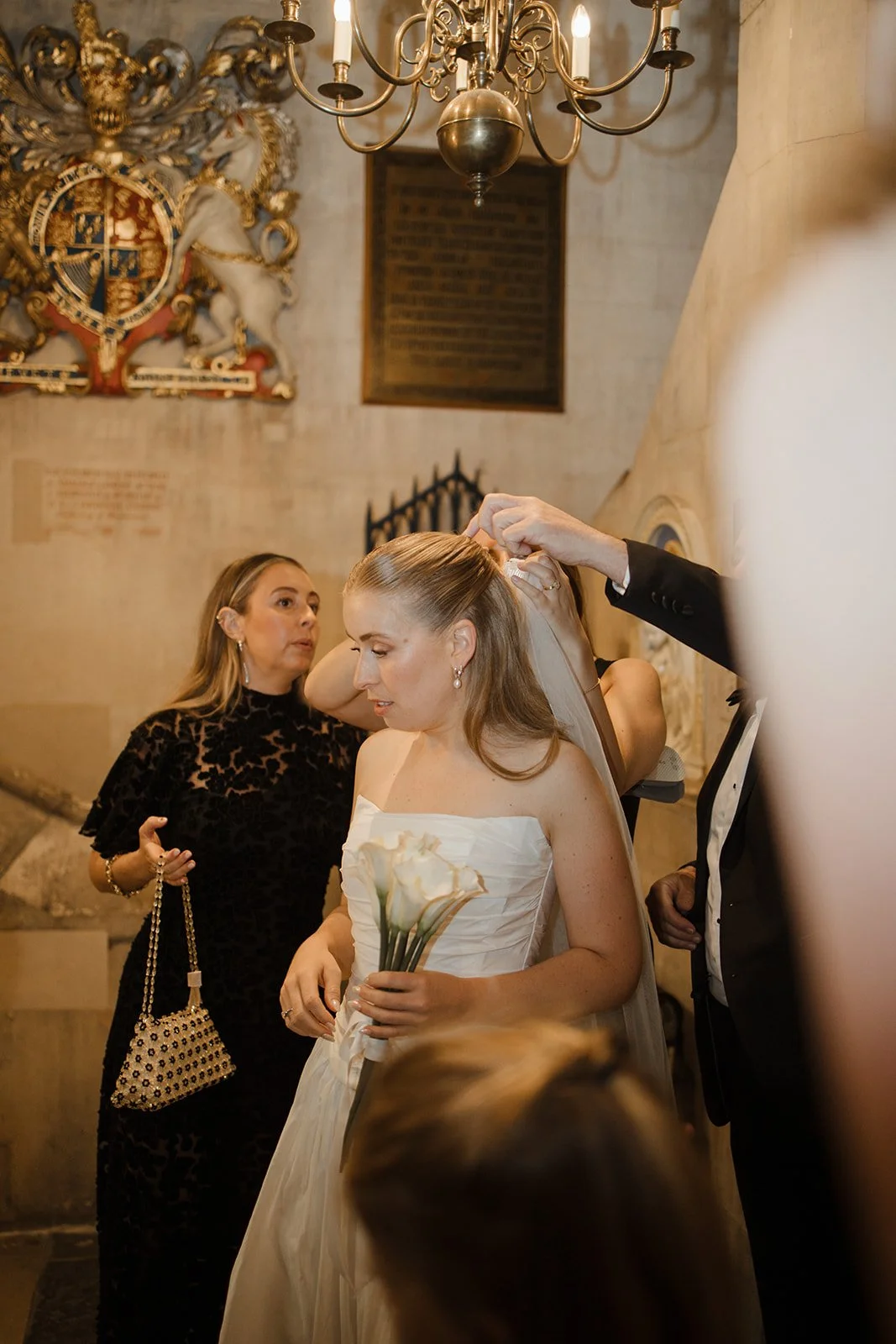 A bride with a bouquet of white calla lilies, getting ready as a woman helps her put on a veil in a church with a chandelier and a coat of arms in the background.