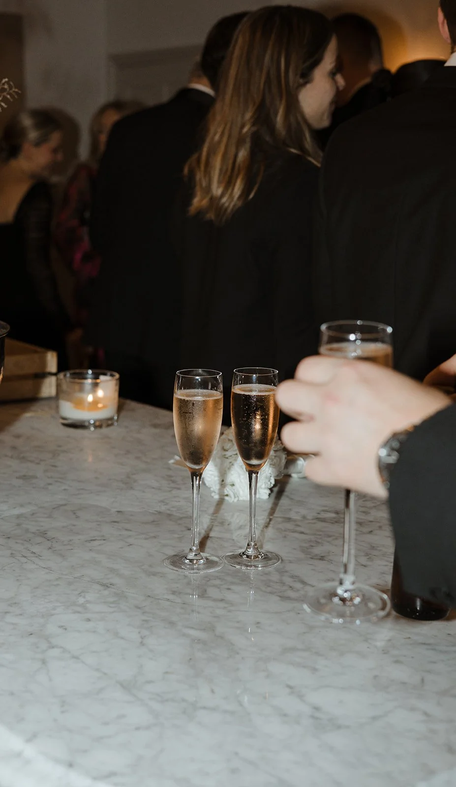 People at a party or celebration holding champagne glasses filled with sparkling wine, with a table featuring a candle, a white flower decoration, and a marble surface.