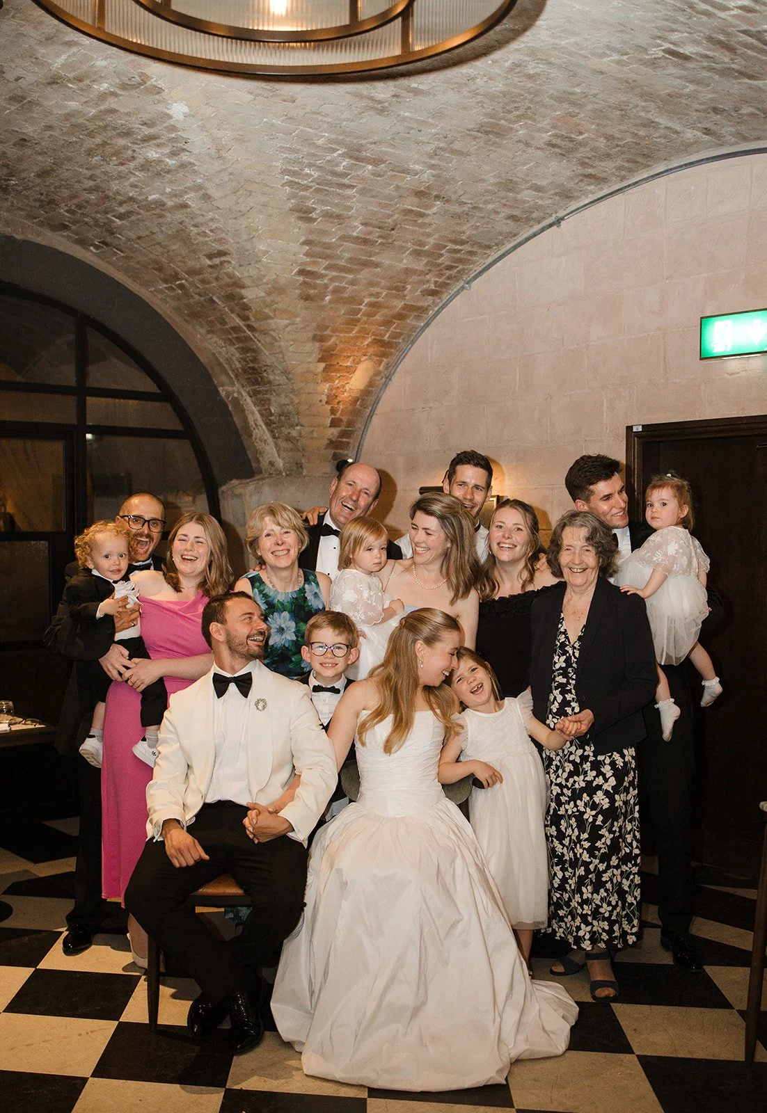 A group of people celebrating at a wedding reception, including children and adults dressed in formal attire, in a venue with a brick ceiling and checkered floor.