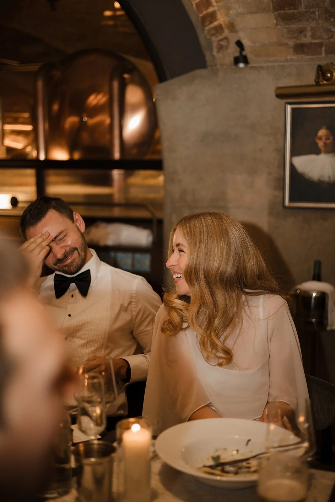 A man and woman are sitting at a dinner table, smiling and laughing. The man has a beard and is wearing a tuxedo with a bow tie. The woman has long, wavy red hair and is wearing a light-colored dress. They are in a warmly lit restaurant with exposed 