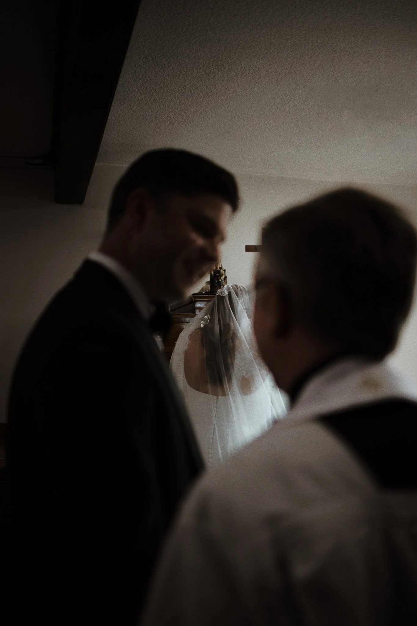 A wedding ceremony with the bride with a veil and two men, one in a tuxedo and the other possibly a priest or officiant, in a dimly lit room.