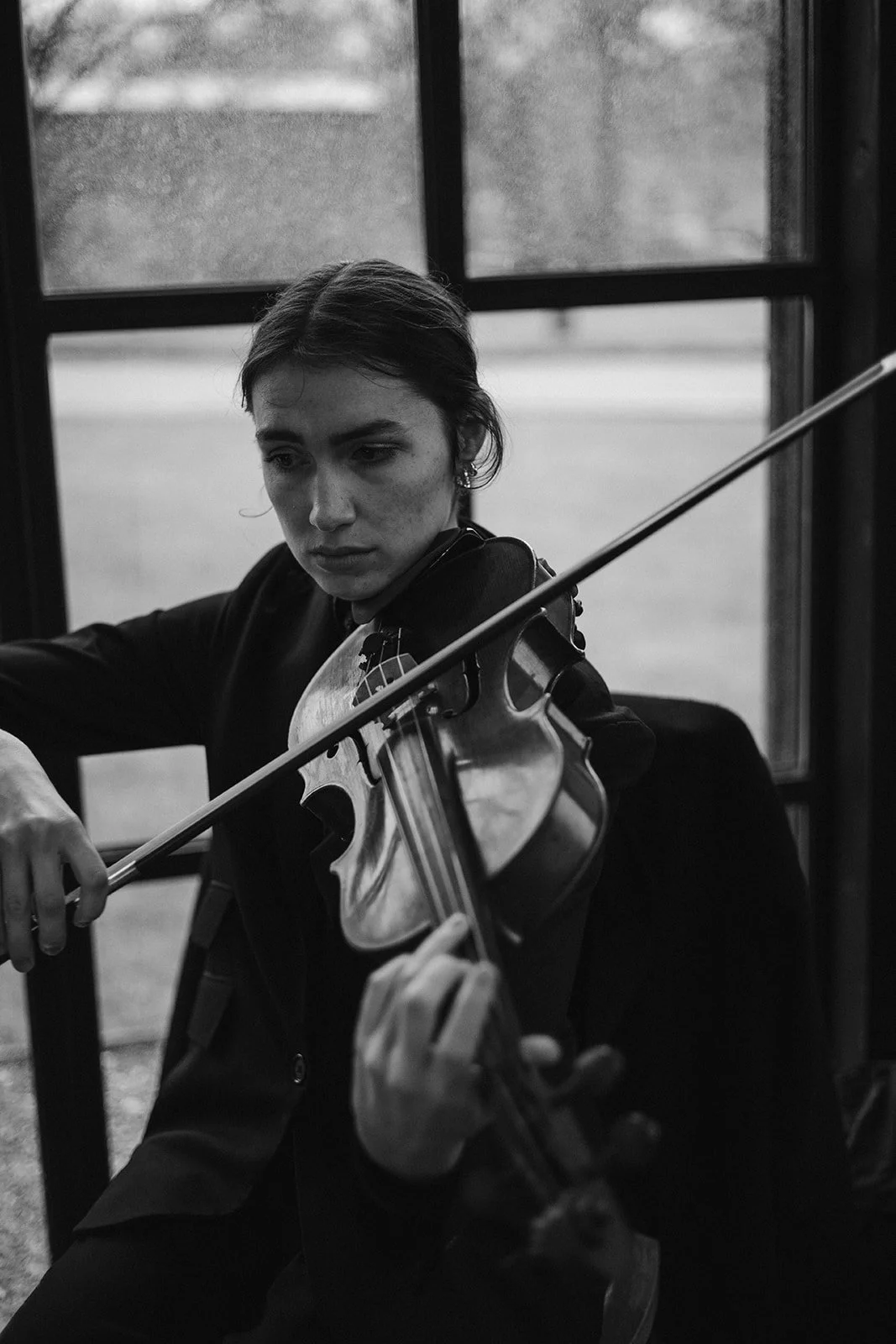 A young woman playing the violin indoors near a window, in black and white.