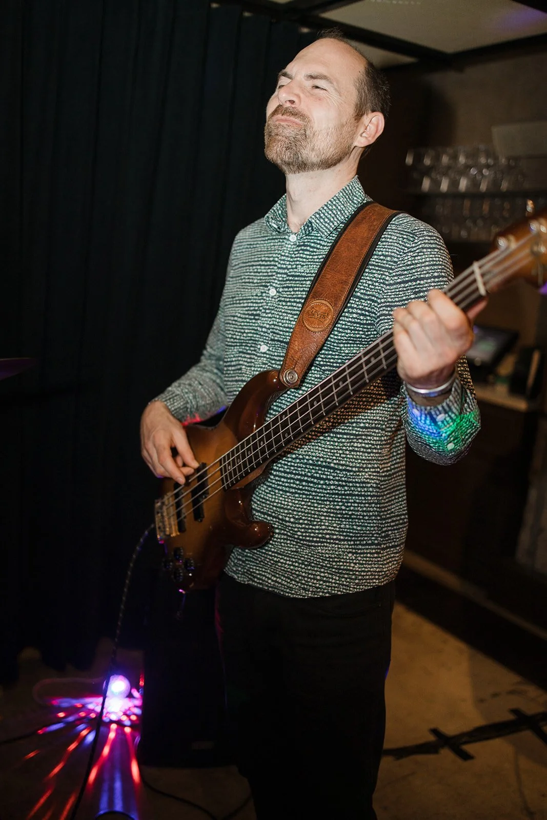 A man with short hair and a beard playing a bass guitar in a dimly lit room, with closed eyes and a joyful expression.