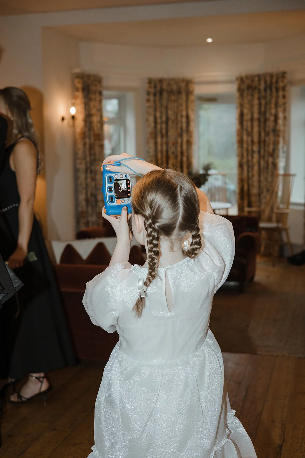 A young girl with braided hair in a white dress taking a photo with a digital camera in a room with wooden floors, armchair, windows, and curtains.