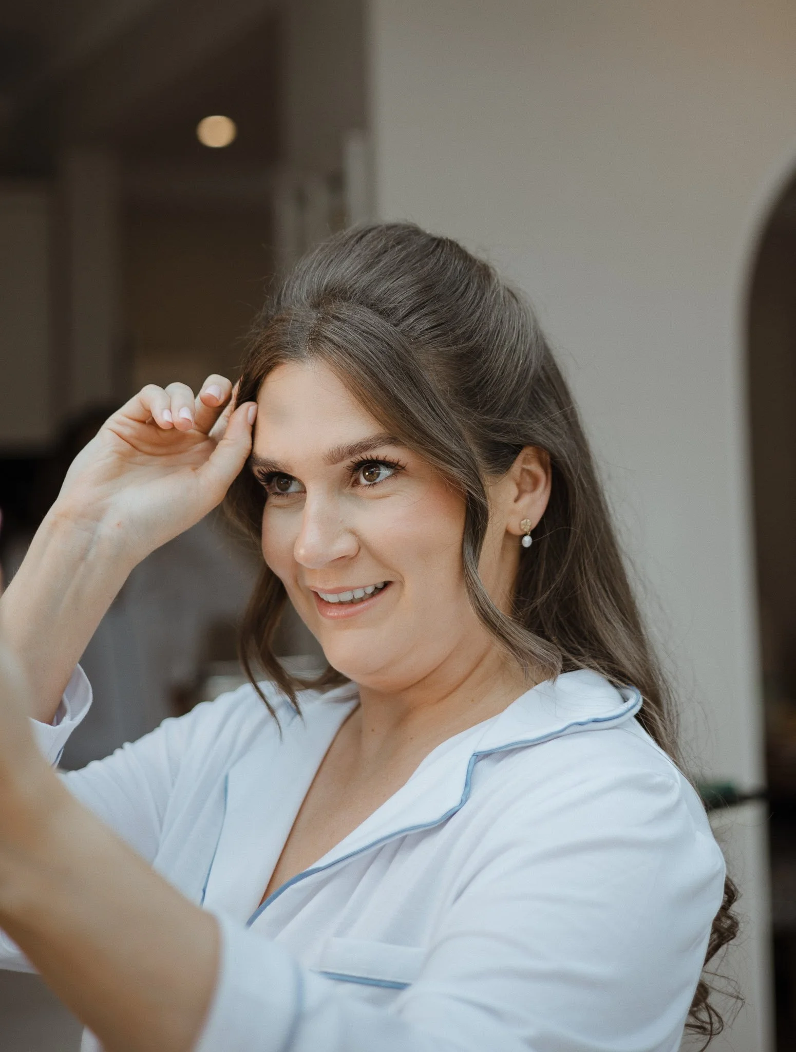 A woman with dark brown hair styled half-up, wearing pearl earrings and a white top, smiling and touching her forehead.