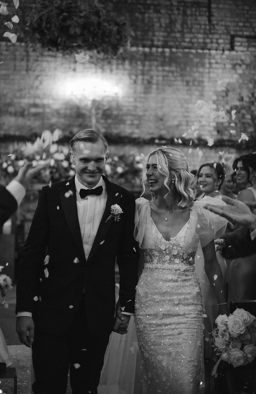 Black and white photo of a newlywed couple walking hand in hand down the aisle, surrounded by smiling guests, with falling flower petals