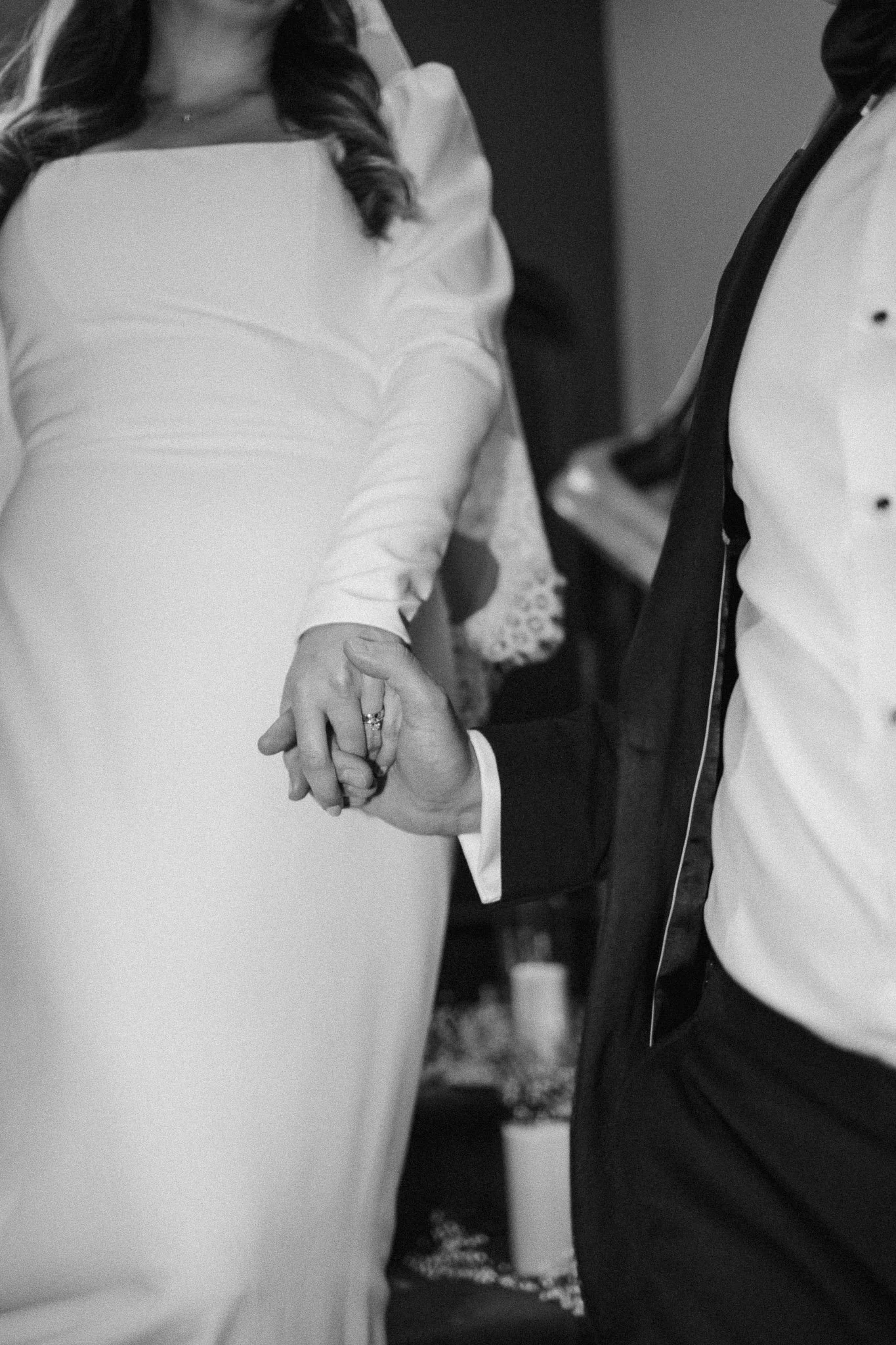 Close-up of a couple holding hands, with the woman wearing a white dress and the man in a tuxedo, during a wedding or formal event.